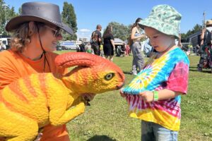 A photograph of a person holding a small dinosaur next to a small child in summer clothes and a sun hat