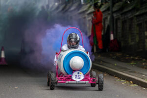 A photograph of a Krazy Races soapbox going down a hill with purple smoke coming out the back