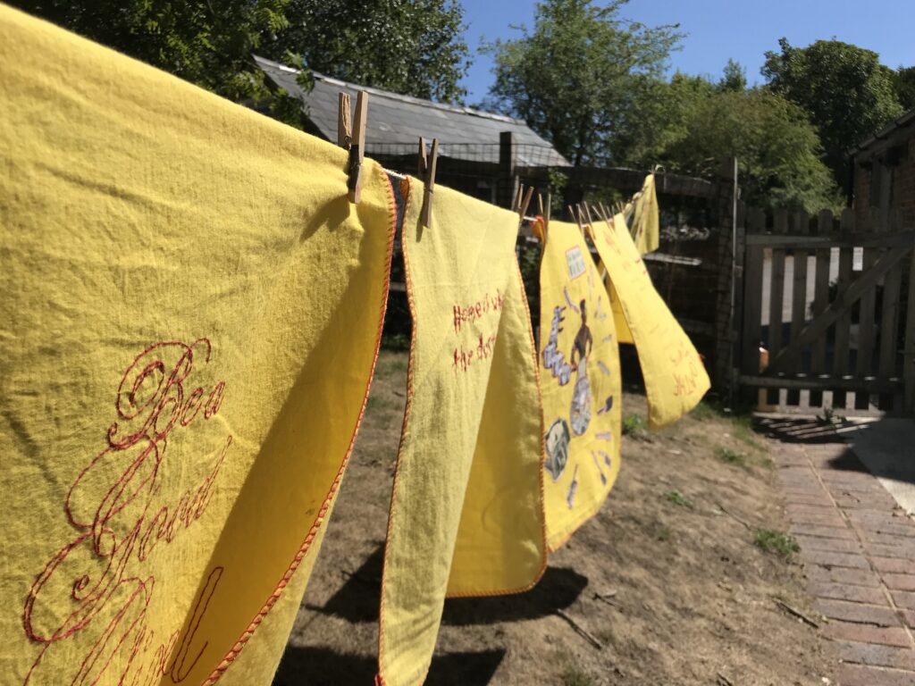 A photograph of embroidered yellow dusters hanging on a washing line in a museum garden in Ditchling