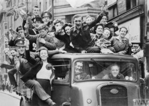 A black and white photograph showing several people riding on a car, waving and celebrating on VE Day in 1945